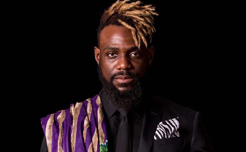 a black person with a beard, a patterned purple shawl over one shoulder, black suit, zebra striped kerchief in pocket, a shock of blond dredlocks spinging from the top of their head
