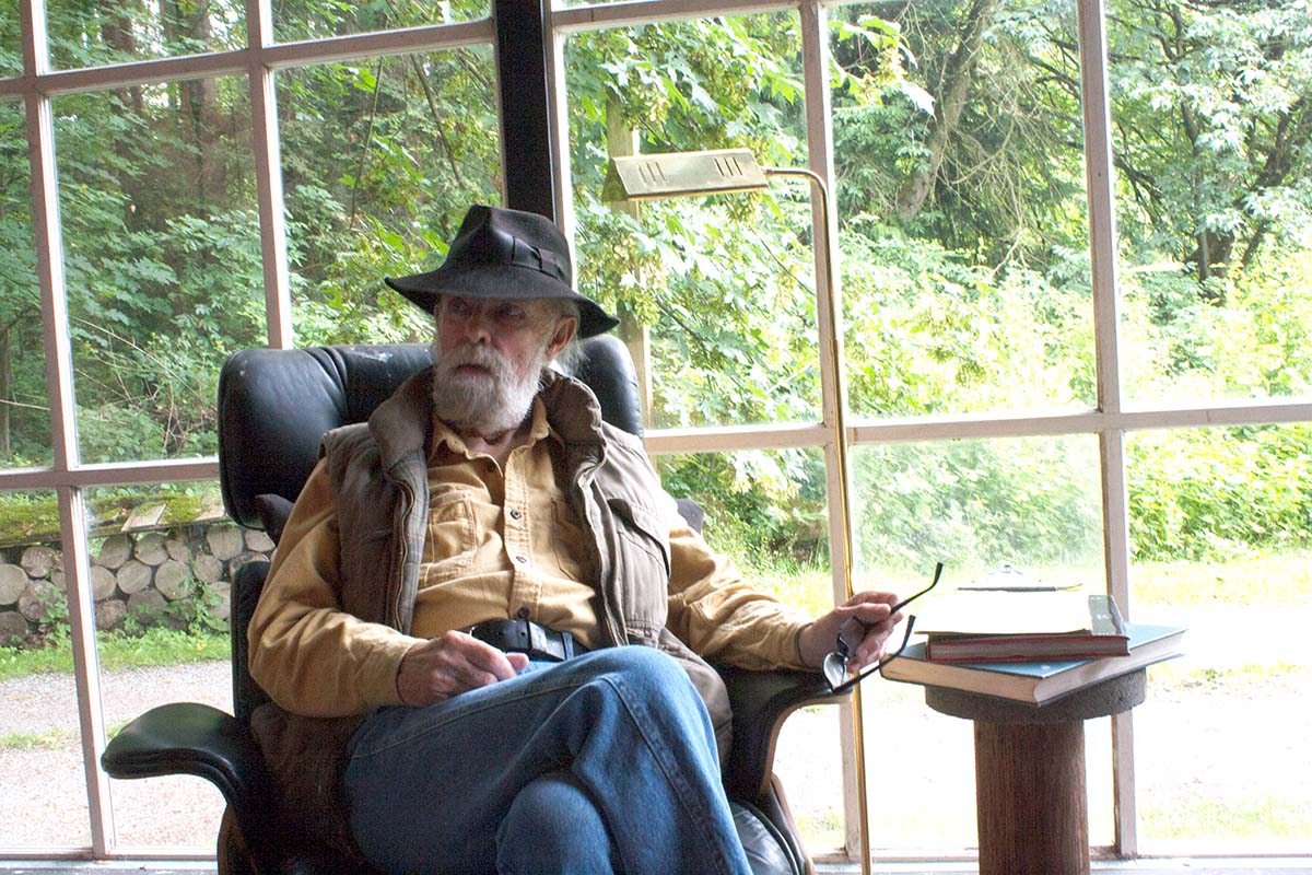 bearded fellow in a brim hat and vest sits in front of a window. outside it is leafy.