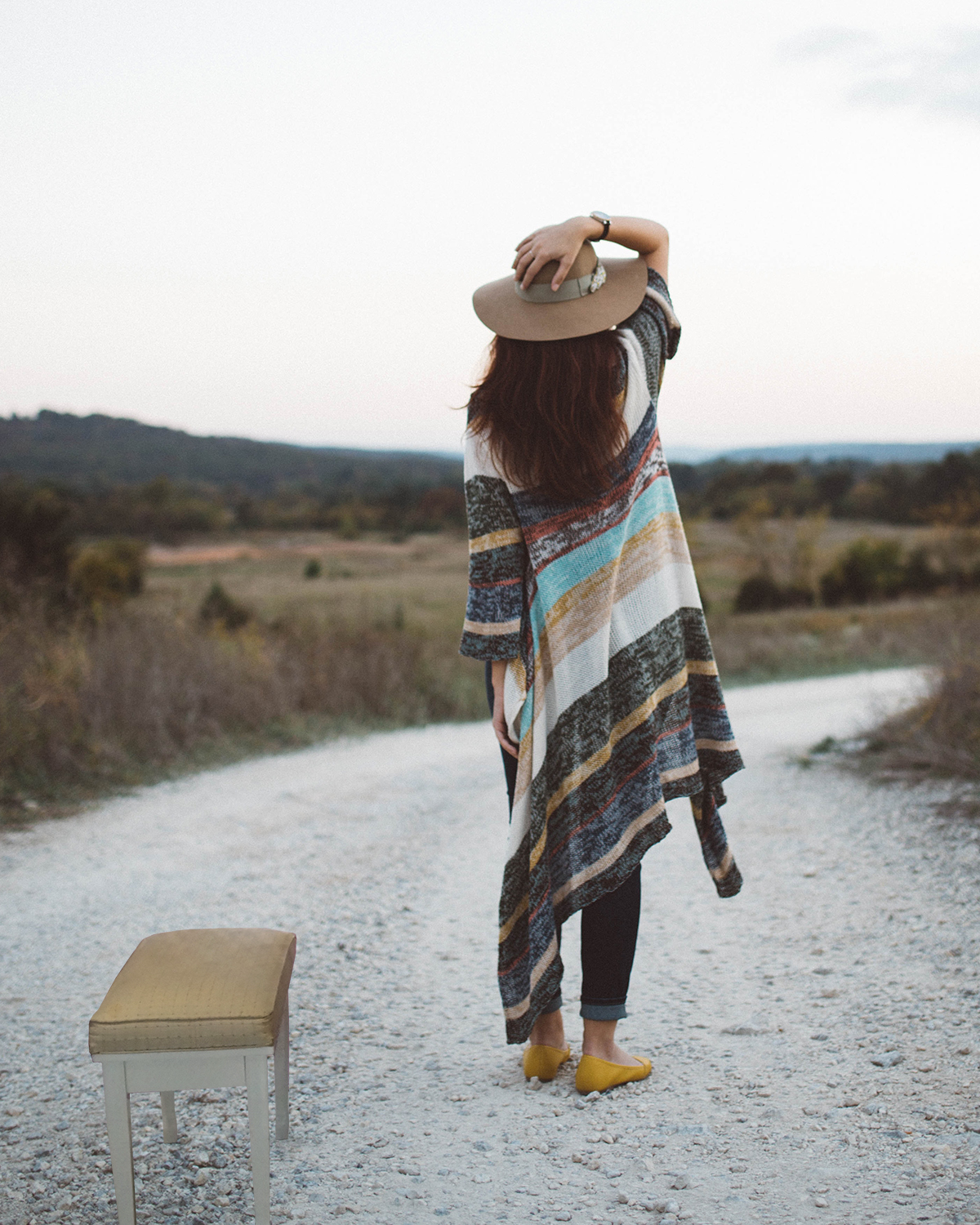 A person with long cardigan and long hair stands staring down a gravel road in the desert, with one hand on their hat on their head. Next to them is a piano bench, also in the middle of the road.