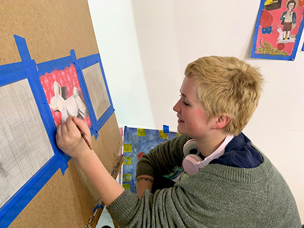 A person smiling as they add detail to an illustration taped to an easel
