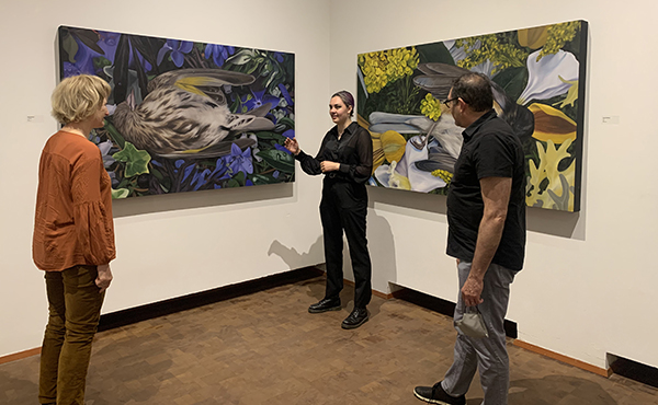 a person standing between two paintings in a gallery, talking with a smile to two other people. The paintings depict birds laying on their backs among flowers.