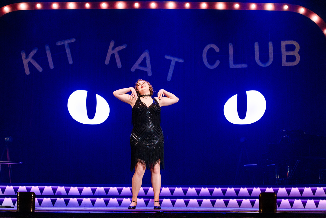 Young club performer emotionally sings in front of glowing cat eyes and a sign for the "Kit Kat Club".