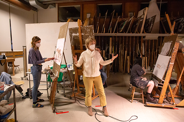 A person speaking, wearing a respirator mask, to a class in an art studio. Students are painting art on easels. In the background is a wall of vertical storage full of paintings.