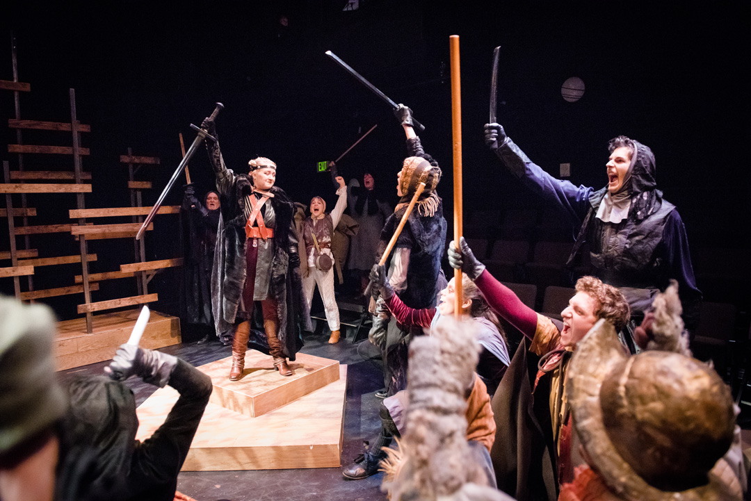 Henry V riles up soldiers in battlewear as they cheer and lift their weapons in solidarity.
