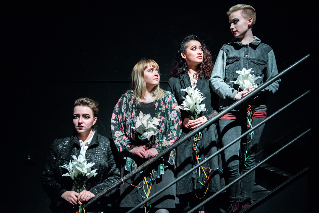 Four actors in grey and black hold white flowers as they stand at different levels on a staircase.