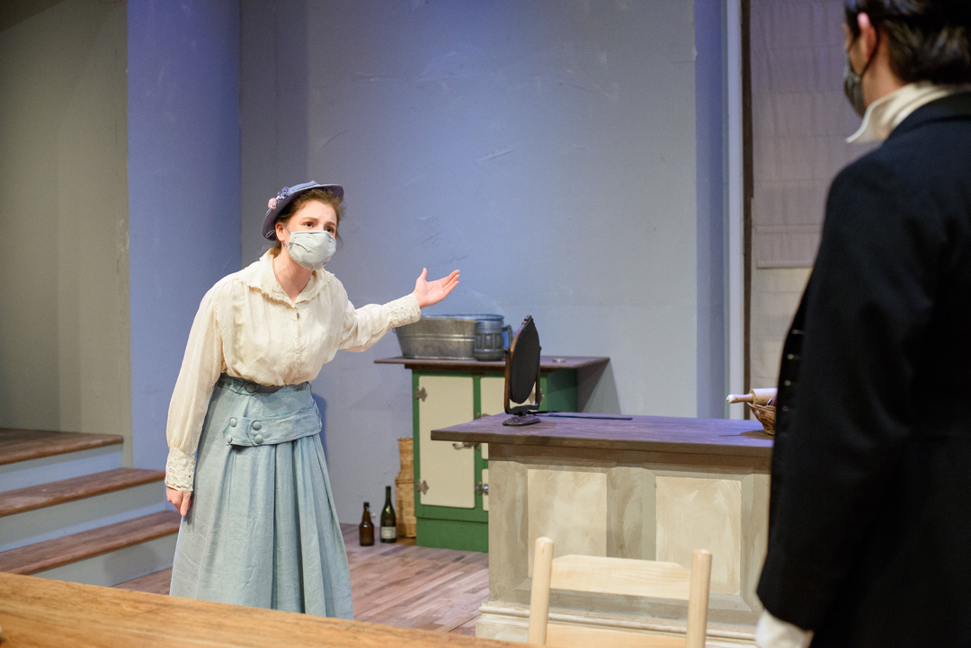 Actor in pale blue skirt and cream blouse gestures in frustration speaking to an actor with their back turned away from the audience.