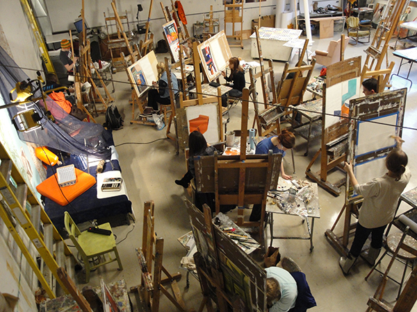 Looking down upon a busy, crowded paint studio: several people painting artwork on easels.