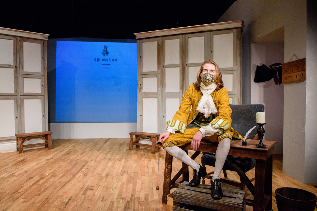 Actor in a frilly, yellow coat and stockings sits atop a writers desk with a quill and candle as the projection behind reads "A Printing House".