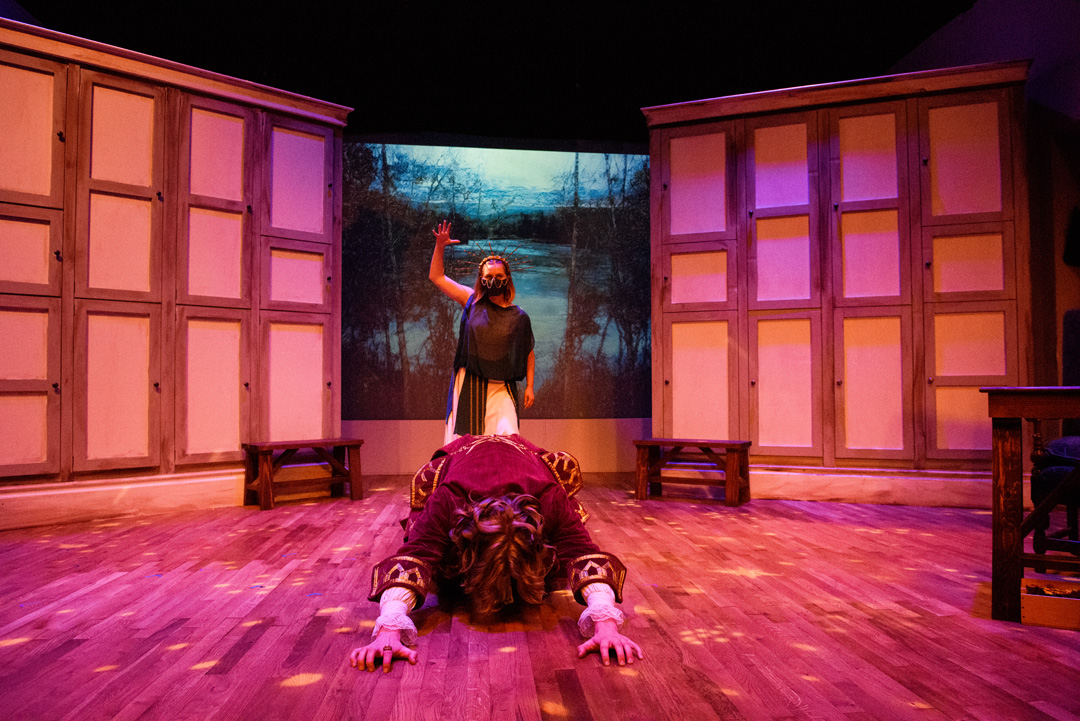 Bathed in red light, a performer with a large crown smites an actor who is bowed on the ground with arms outstretched. 