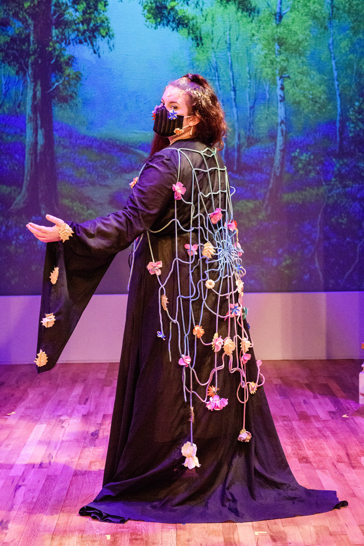 Actor in a long black dress with flowers and a spiderweb draped like a cape looks to the audience and summons to follow.