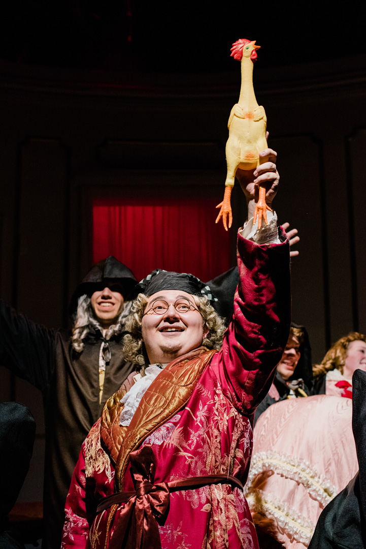 Actor in red and gold robe holds a plastic chicken in the air as performers surround and cheer. 