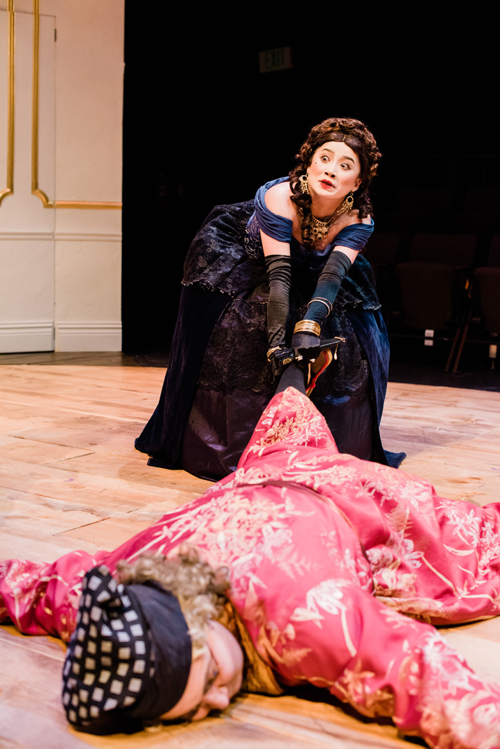 Actor in a ballgown drags another seemingly unconscious and looks up with a surprised expression.