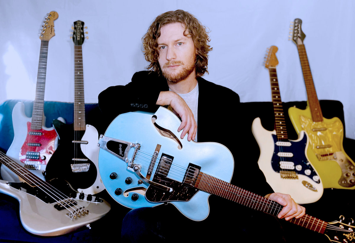 Brendan Byrnes holding a guitar, surrounded by more guitars, looking cool
