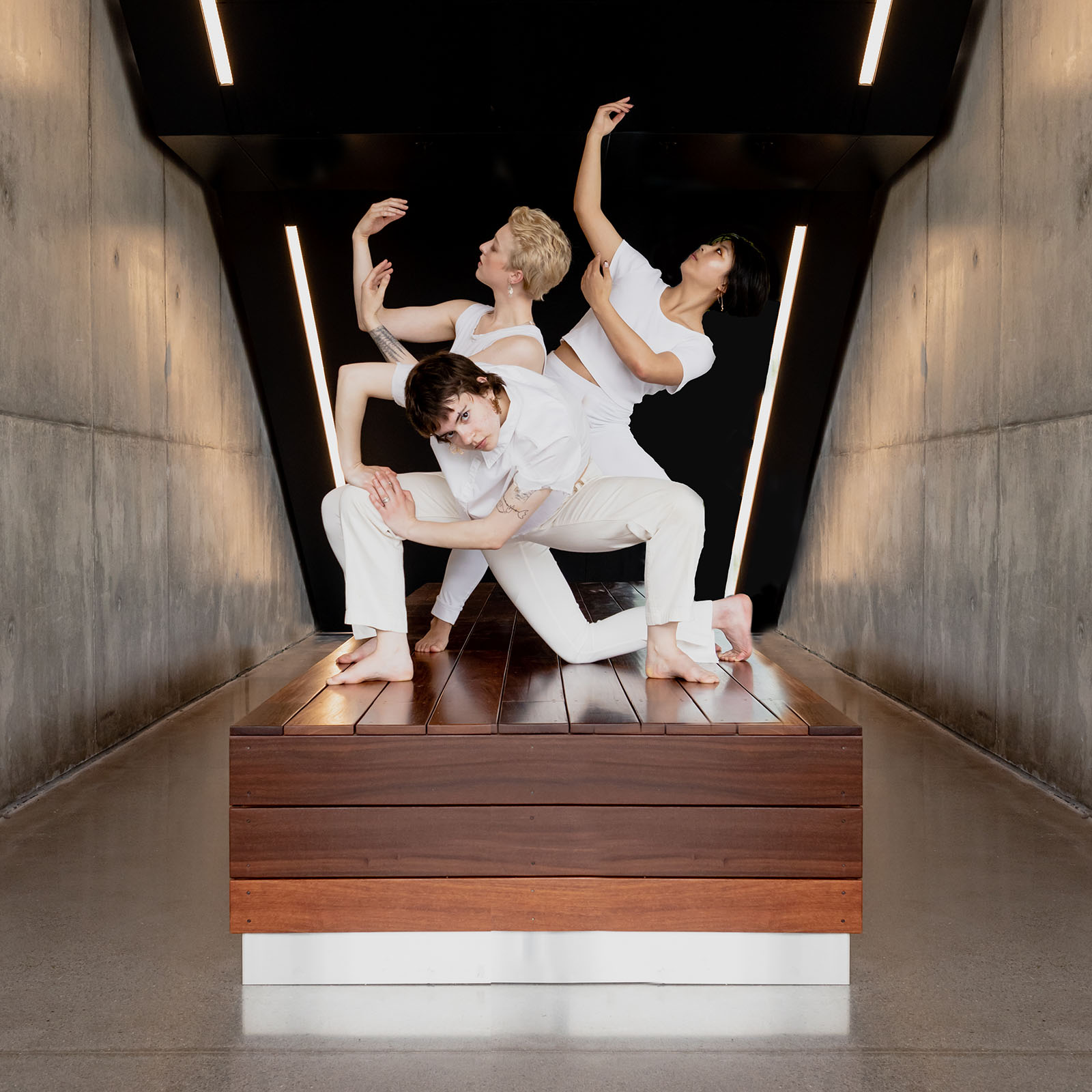 three dancers in white strike low, angular poses in a small space of concrete and wood with modern lighting and details