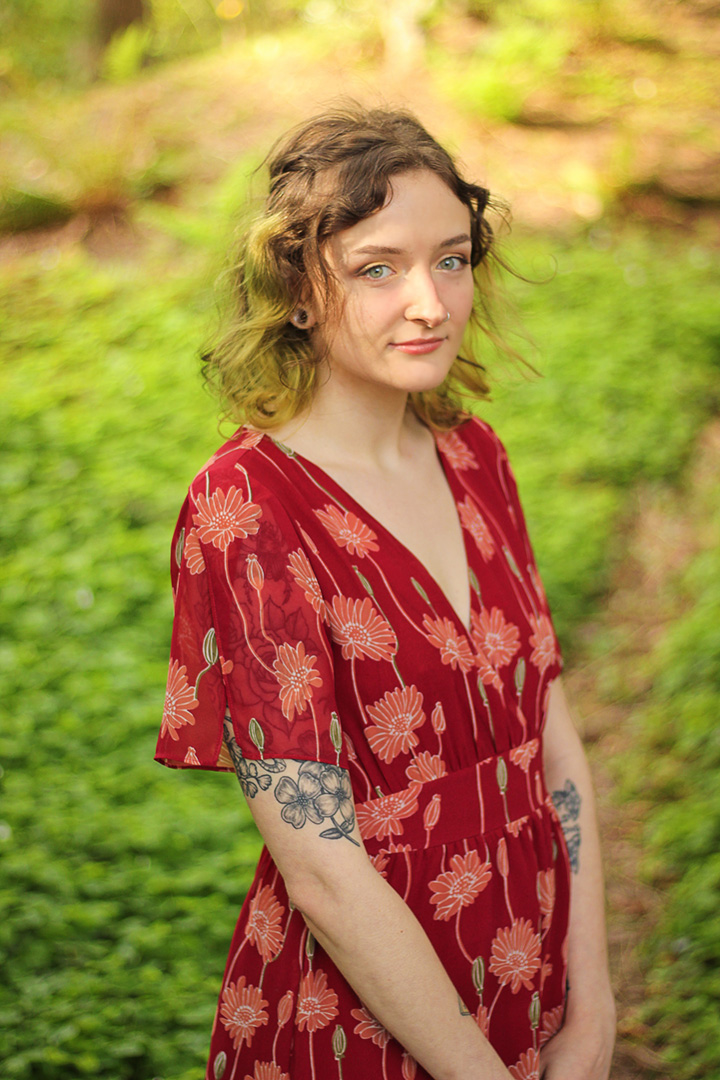 Emily Castle standing with a subtle smile. Emily has flowery tattoos on both arms