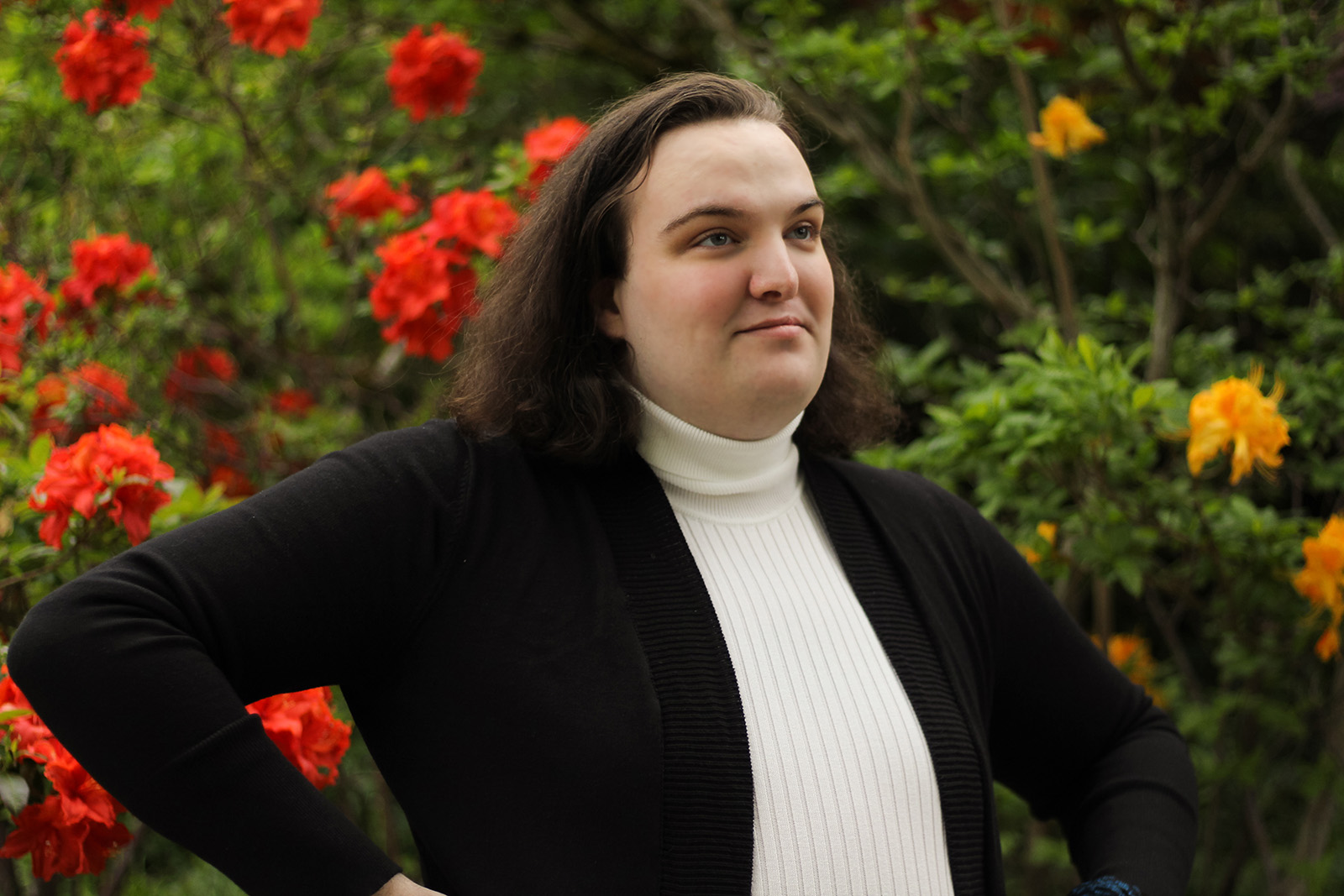 a person in a white turtleneck and black blazer stands in front of flowering shrubs