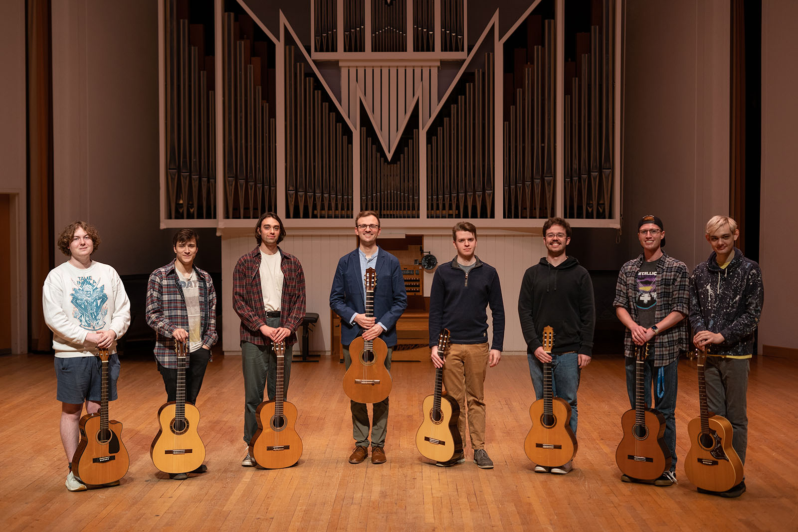 guitar players stand on a stage in a semicircle holding their guitars. There is a pipe organ behind them.