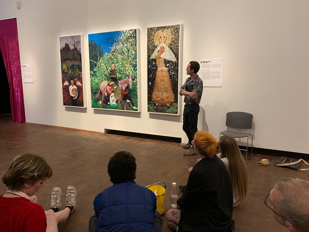 a person stands next to elaborate paintings depicting people and nature, in front of a group of people sitting on the floor