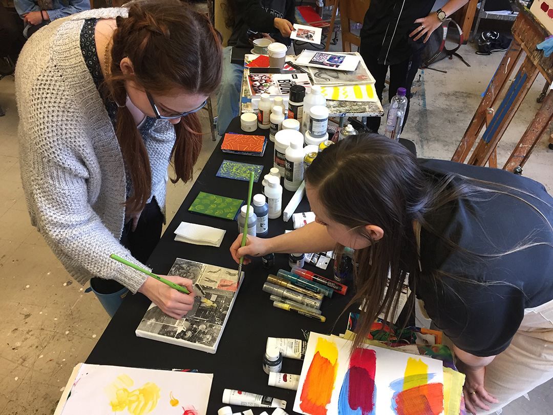 two people painting on the same canvas, on a table filled with art and art supplies