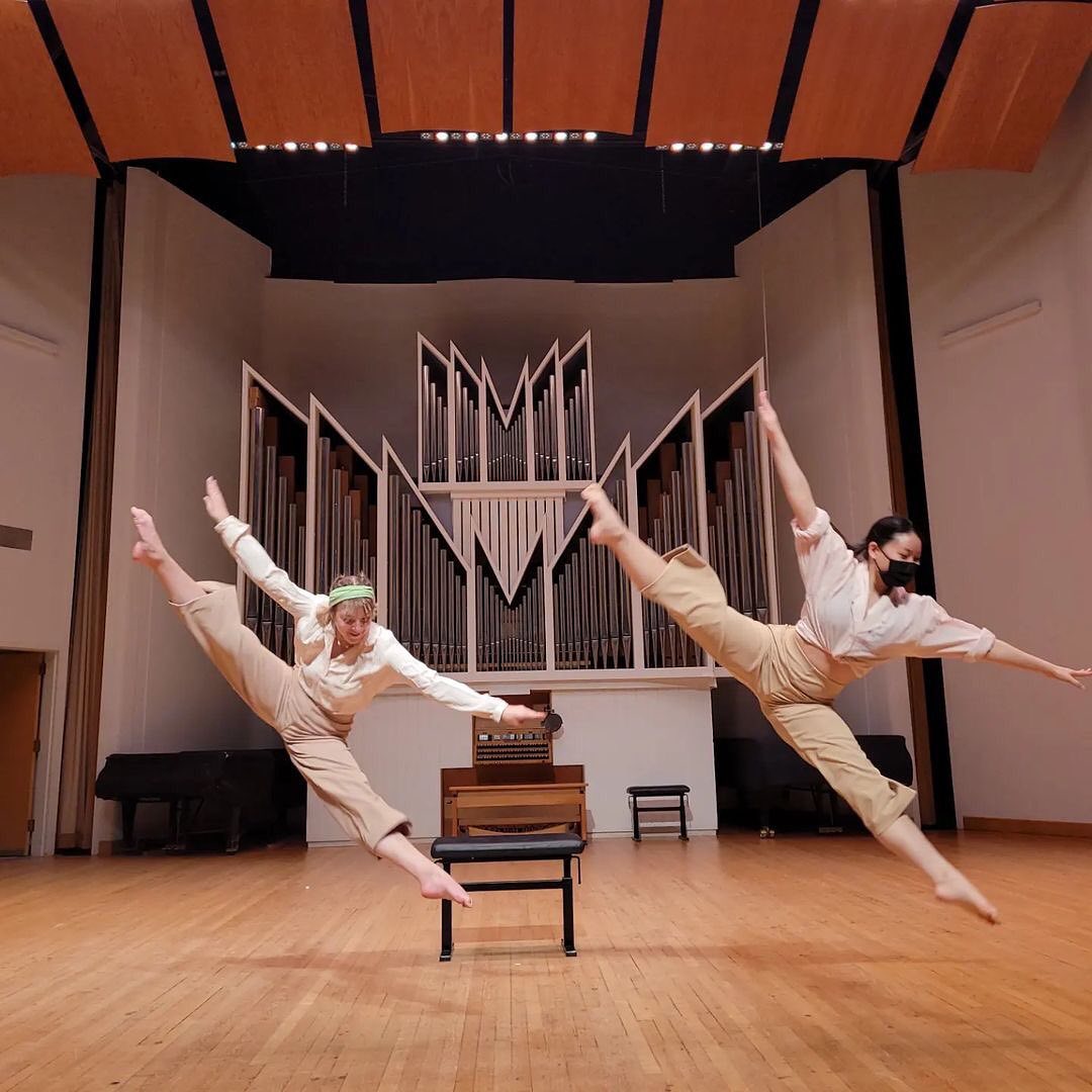 two dancers leap side by side in the splits with their legs at 45-degree angles to the stage. Behind is a pipe organ.