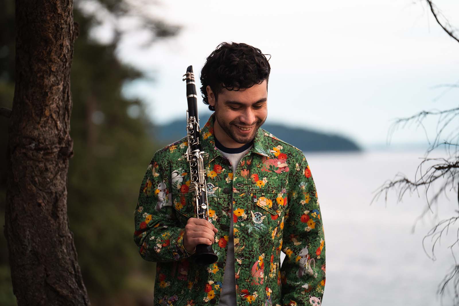 curly-haired person in a floral shirt with a clarinet over their shoulder.  A lake is behind.