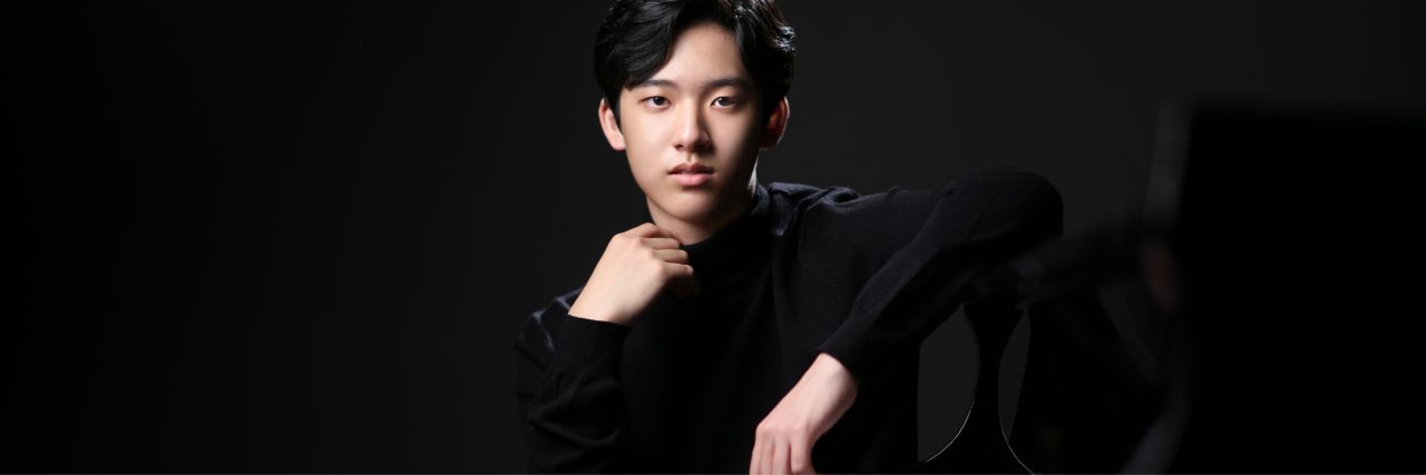 Yunchan Lim, straight faced, leaning an elbow casually on a piano with one hand on his collar