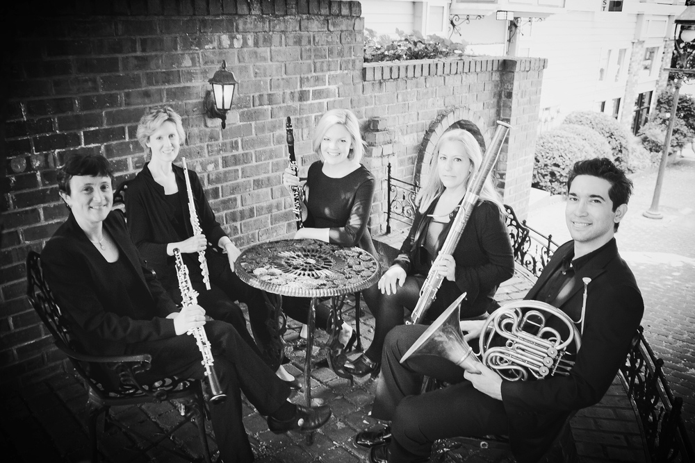 a woodwins quintet seated at an outdoor cafe. They are holding their instruments.