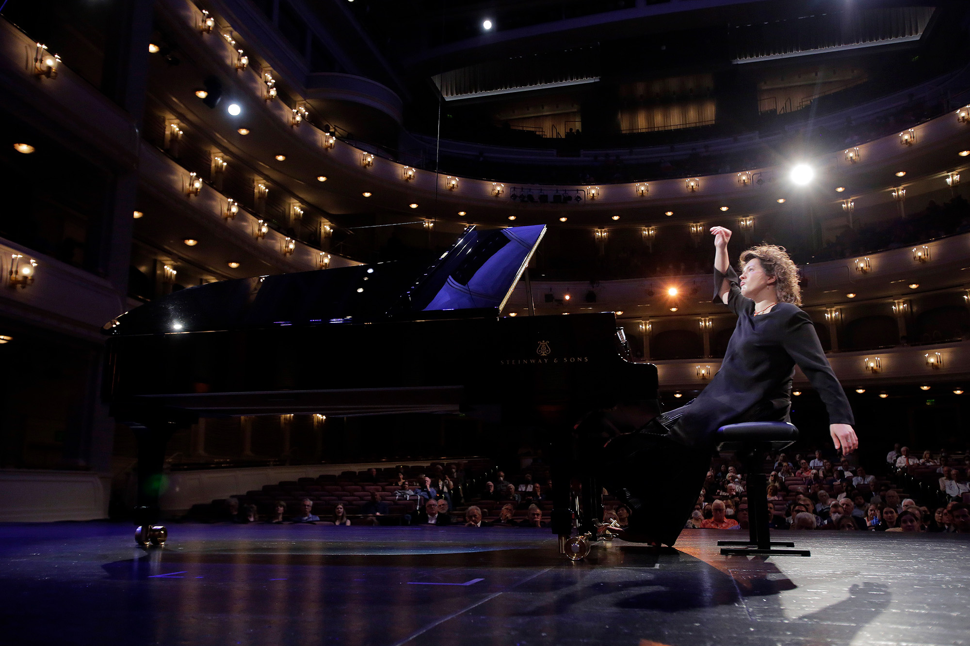 Anna Geniushene seated at the piano in a crowded concert hall, swaying back with a look of focus: one hand swings behind her and the other swings above her head