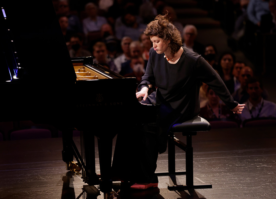 Anna Geniushene playing piano in front of a crowd. Her hair flies as she leans to the side with puckered lips and one hand swinging behind her