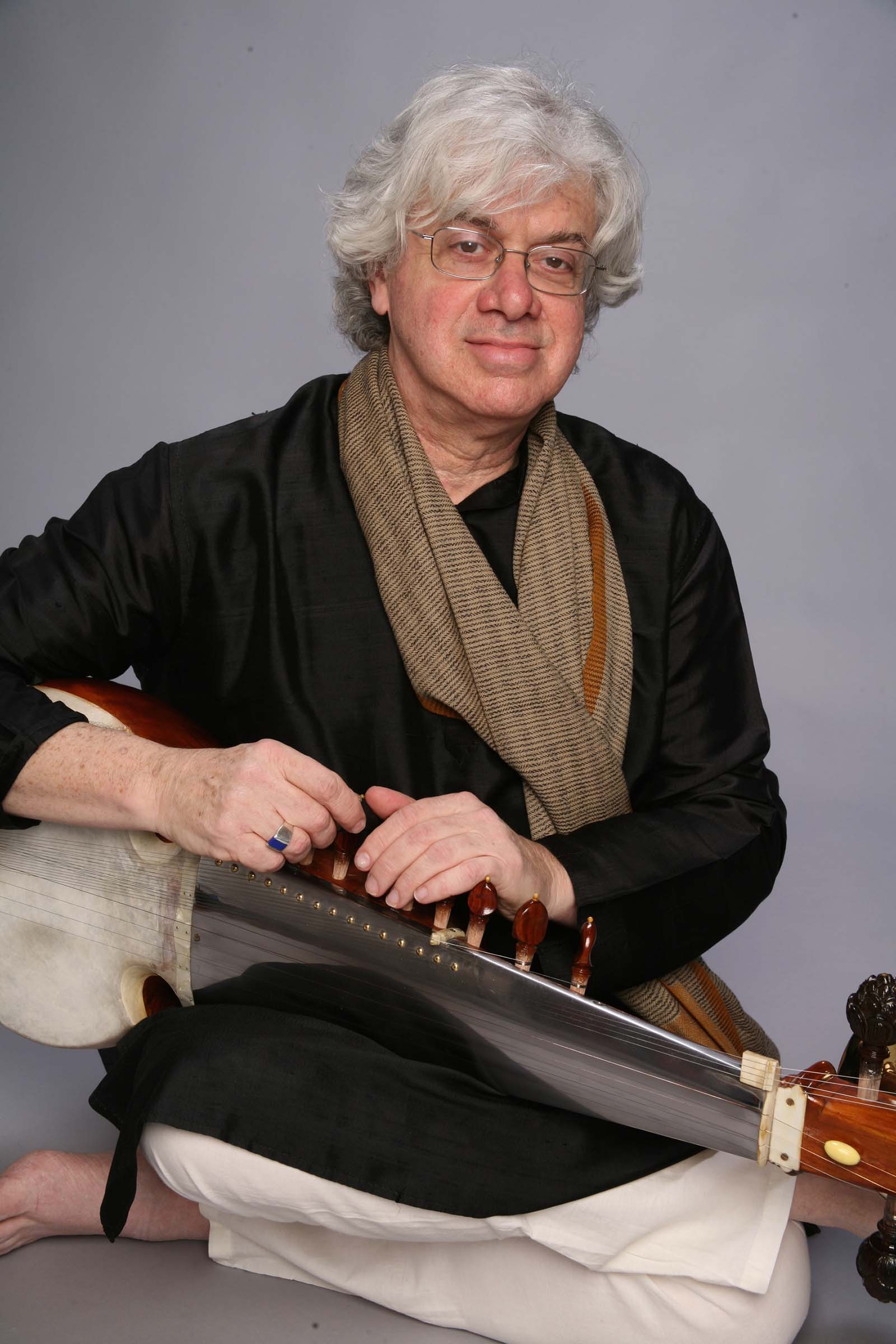 David Trasoff is seated cross-legged on the floor with a sarode across his lap. A sarode is a multi-stringed instrument. David has wavy silver hair and glasses.