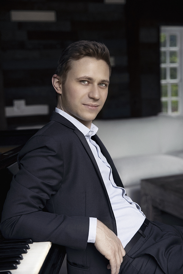 Dmytro Choni in a suit, seated backward at a piano, leaning casually on the keys. Dymtro has an intense gaze and barely noticeable smile.