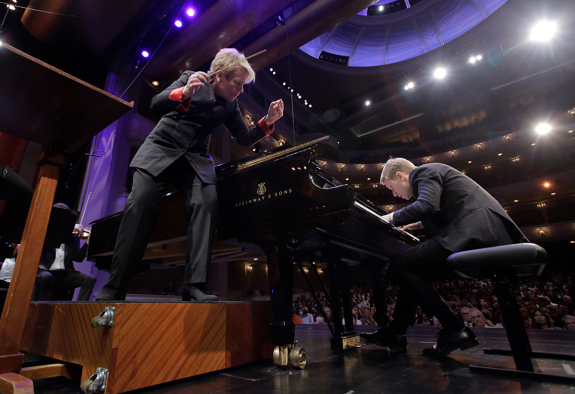 Dmytro hunches intensely over the keys of a piano in a crowded concert hall as the conductor in action peers around the piano to look