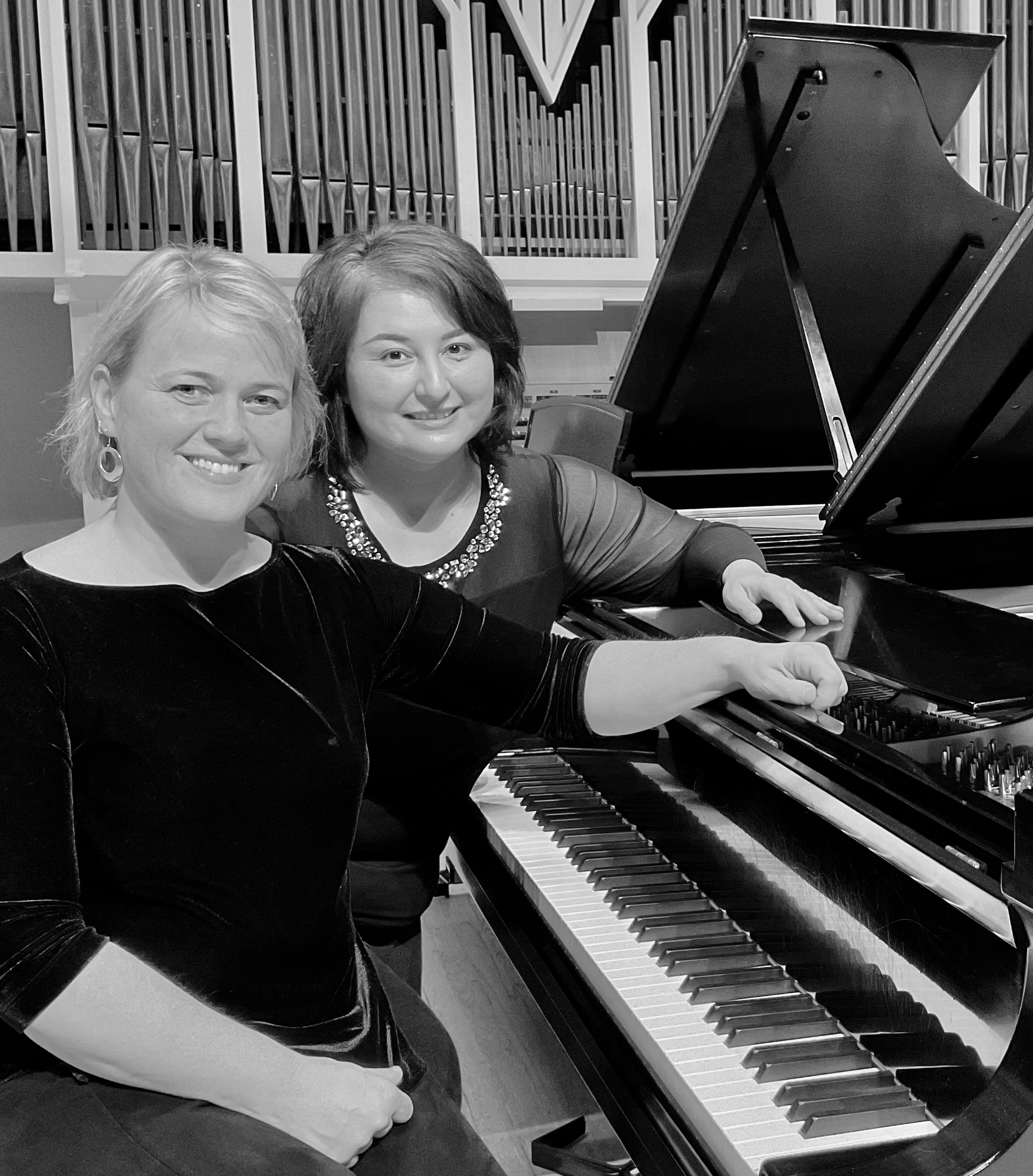Kay Zavislak and Jensina Oliver sitting at a piano together, turned to the side with one hand on the piano top, smiling