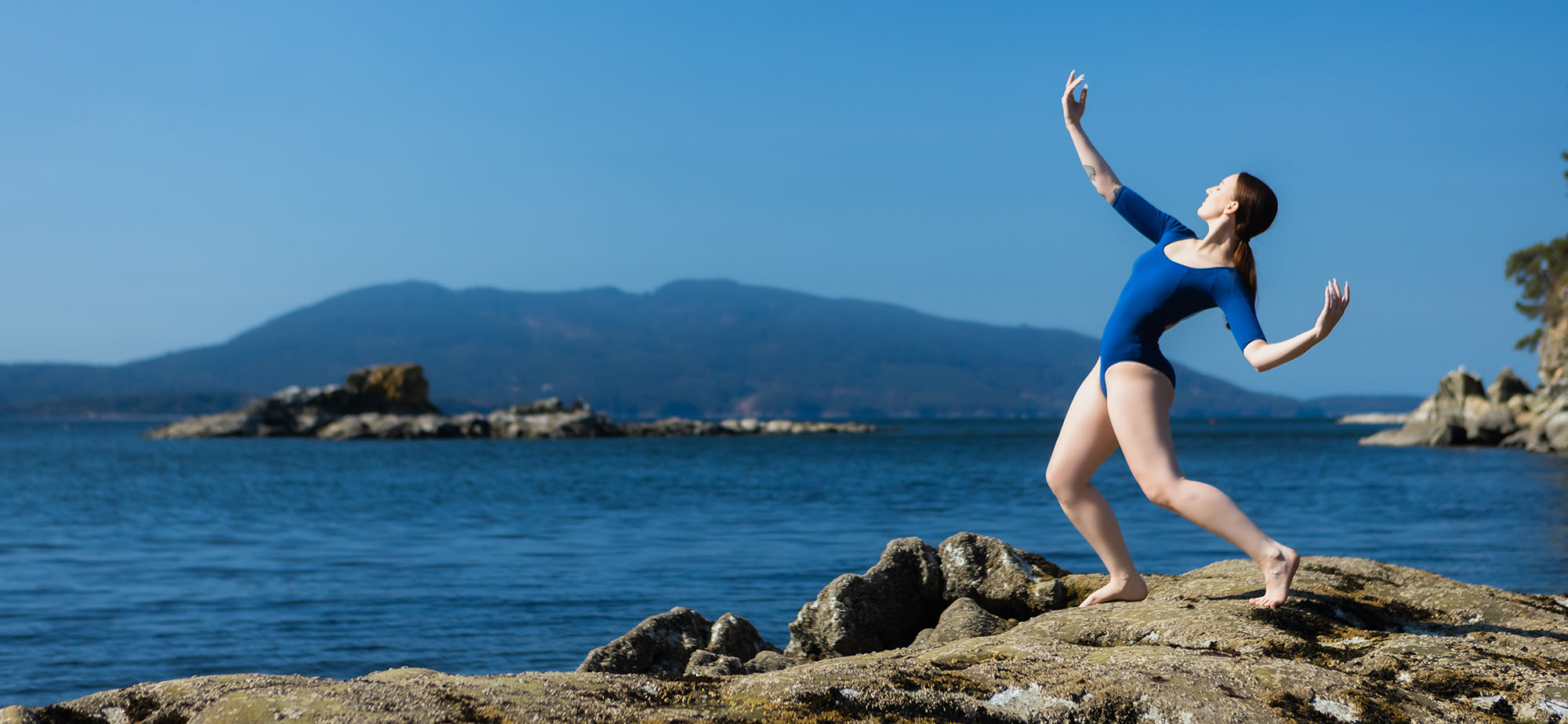 A dancer in a blue leotard stands on a rock by the sea leaning back with arms in a heroic position