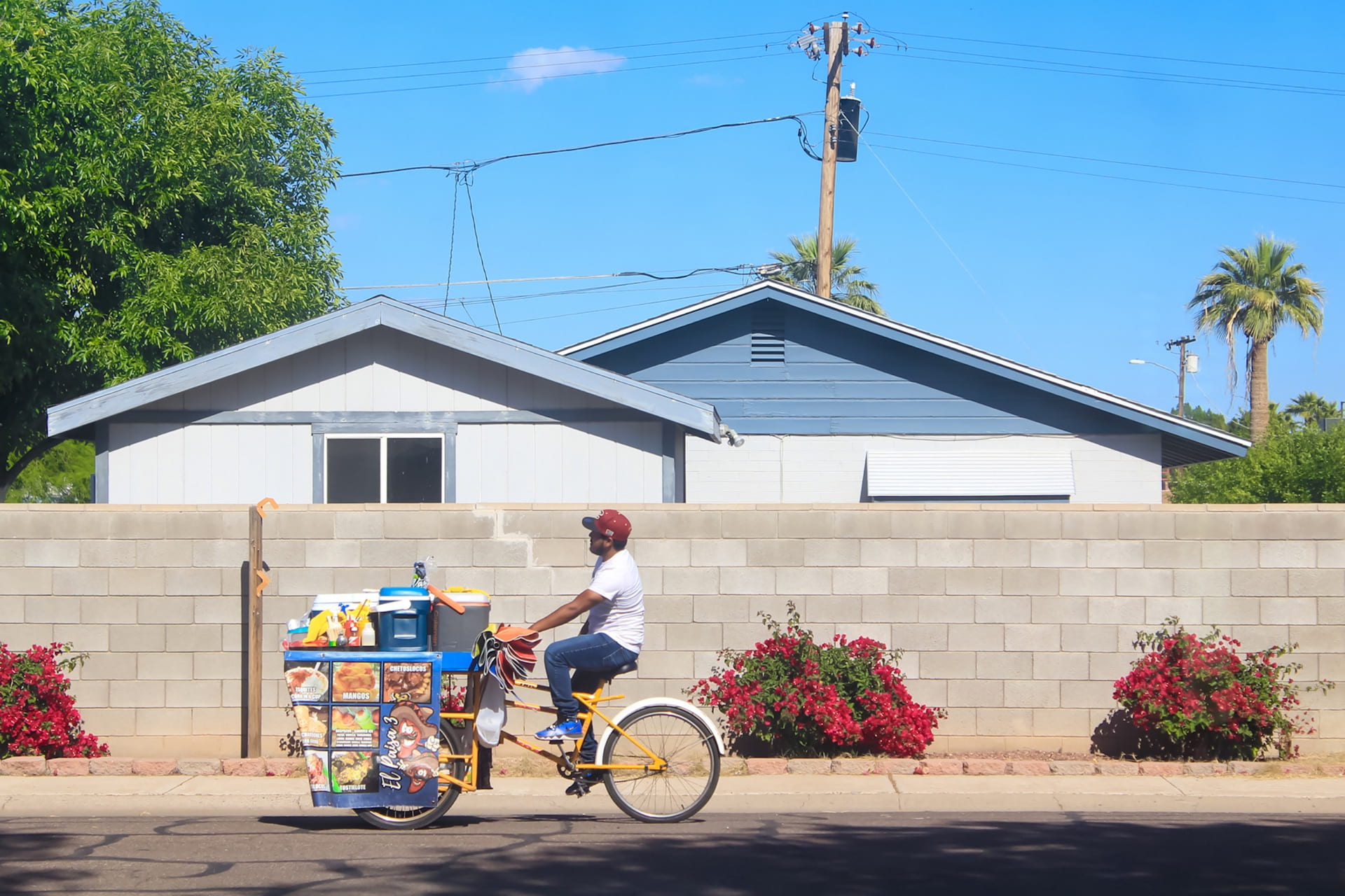 A person cycling past a house with a full cart on the front of the bike, almost as big as the bike