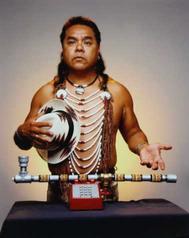 A Native American in traditional attire stands behind an antique telephone - the earpiece replaced by a long pipe.