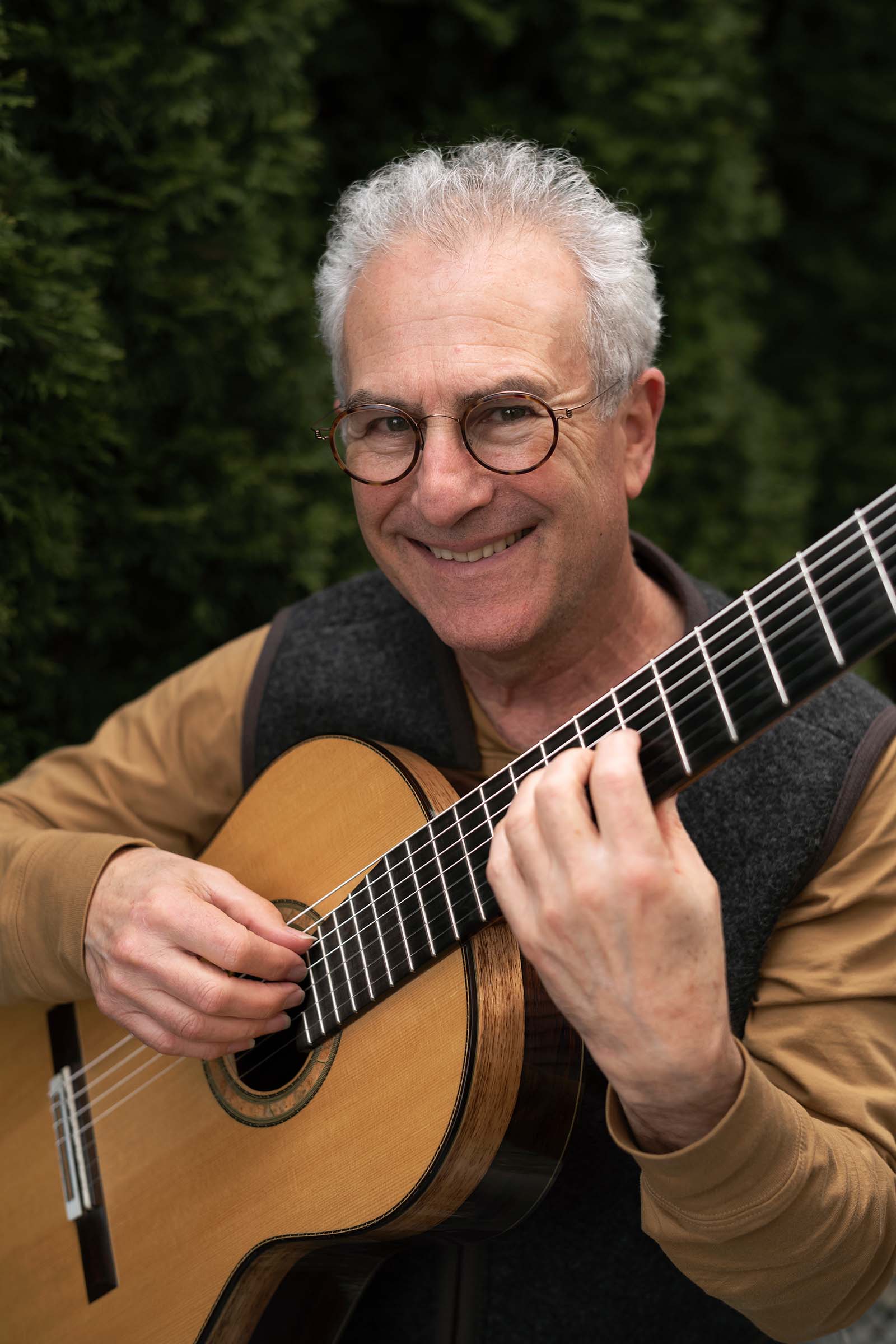 a smiling gray-haired person in glasses holds a guitar ready to play