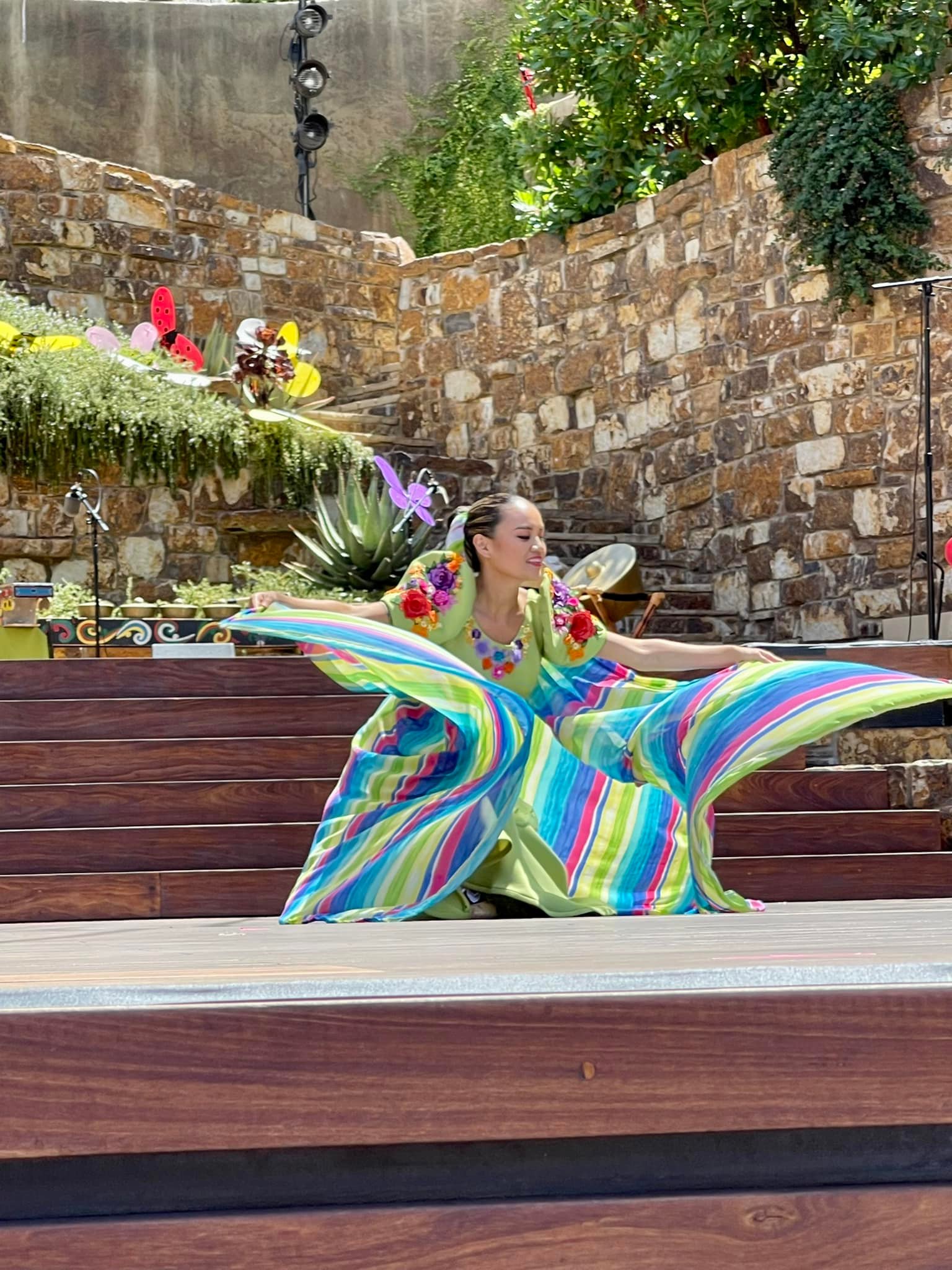 a hula leader makes a broad low gesture with both arms fanning a brightly covered piece of fabric