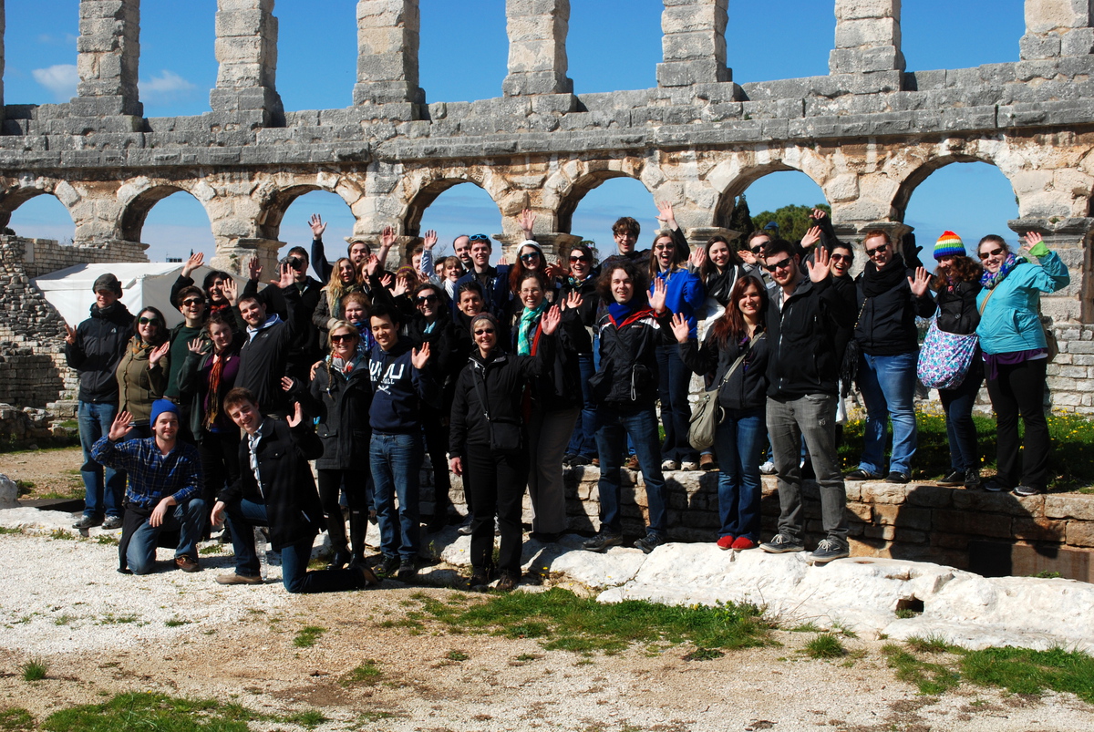 A big group of people in coats smiling and waving outside in front of a multistory stone wall full of arches with blue sky behind