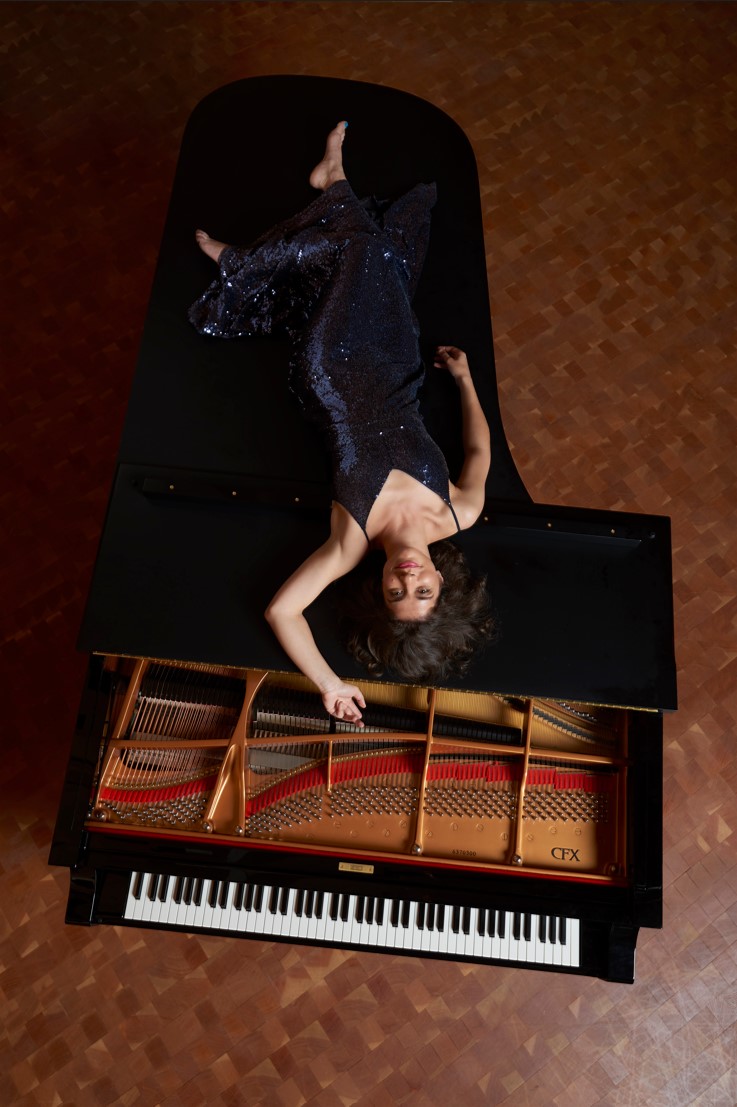 upside-down view from overhead: Inna Faliks in a dark sequined dress, laying on top of a steinway piano, smiling