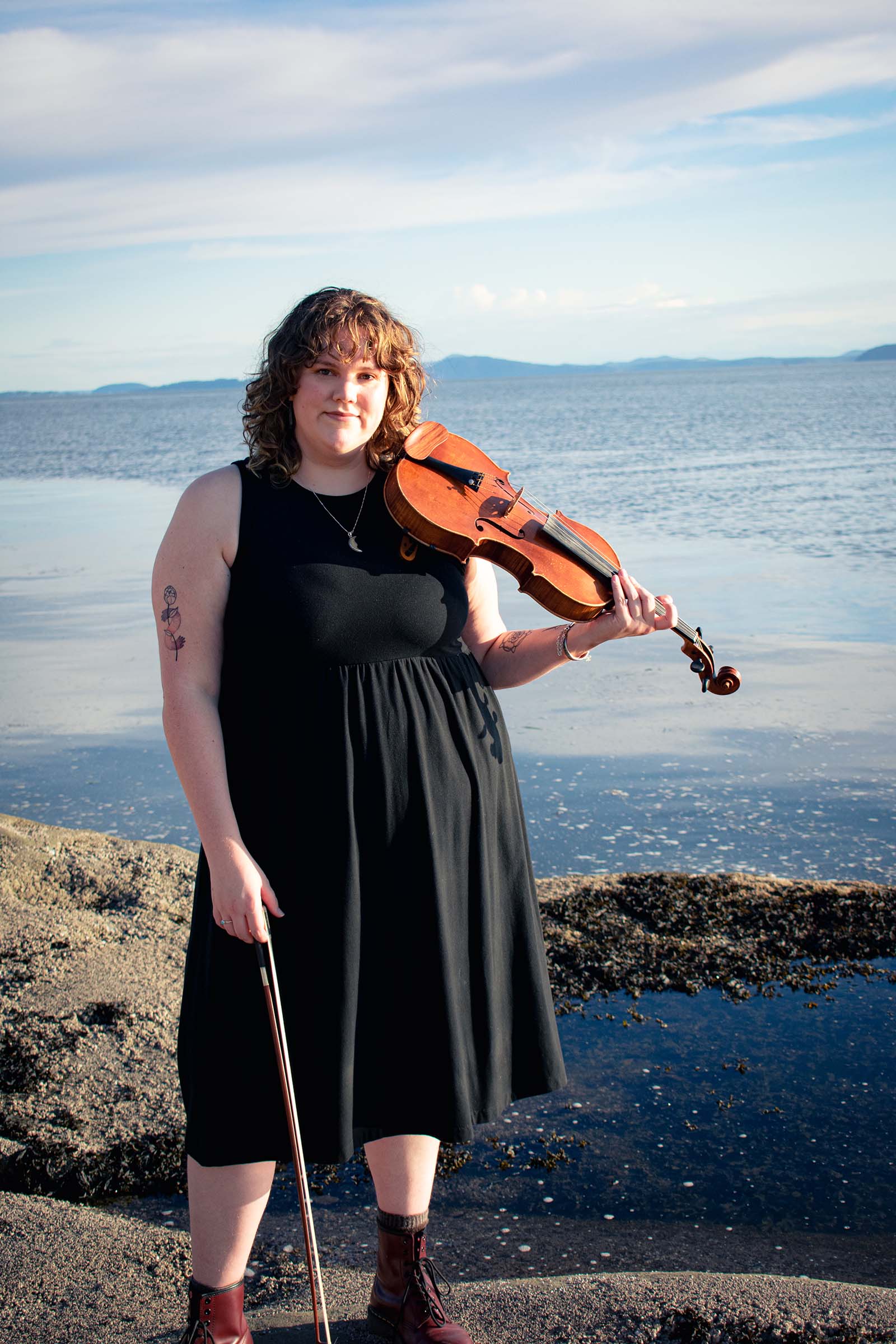 a person in a black sleeveless dress stands at the shore holding a viola