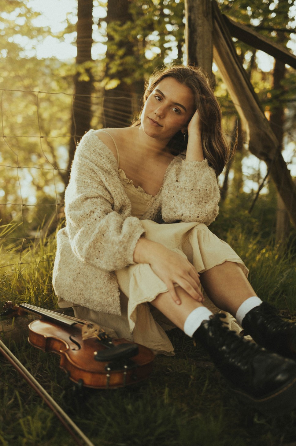 A person sitting peacefully on the grass next to a violin, and against a fence in a warm, woodsy setting