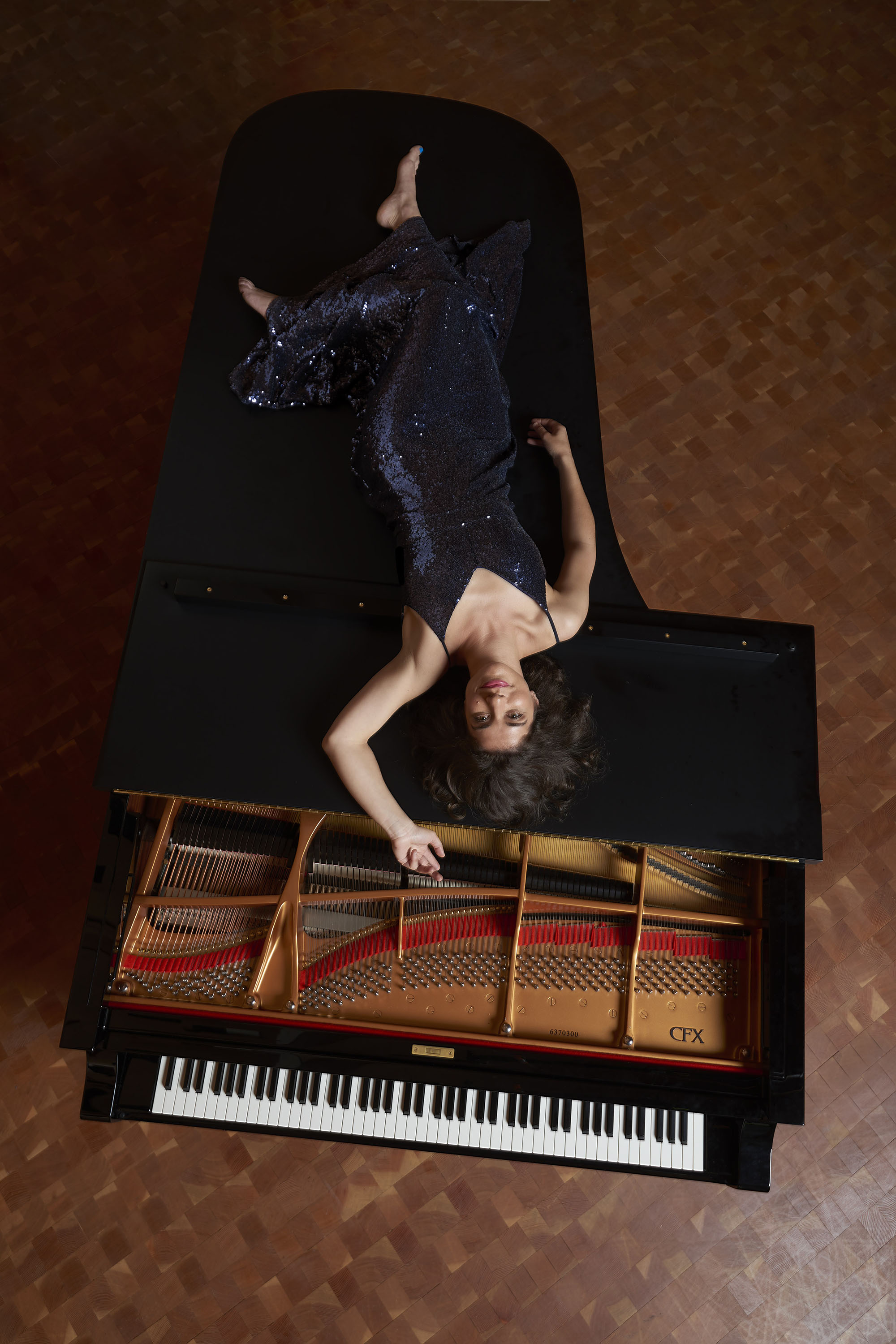 upside-down view from overhead: Inna Faliks in a dark sequined dress, laying on top of a Yamaha grand piano, smiling