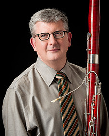 Paul Rafanelli, a person with gray hair, glasses and a business shirt with tie, holds a bassoon
