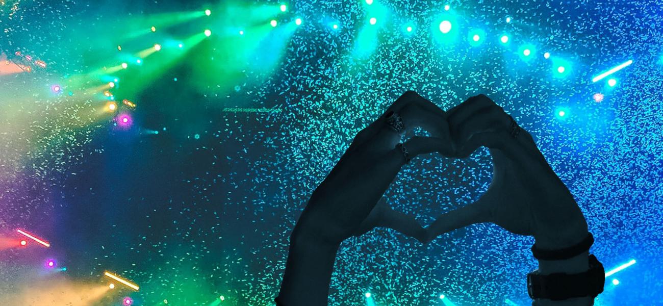 silhouetted hands forming a heart shape in front of colorful stage lights and confetti