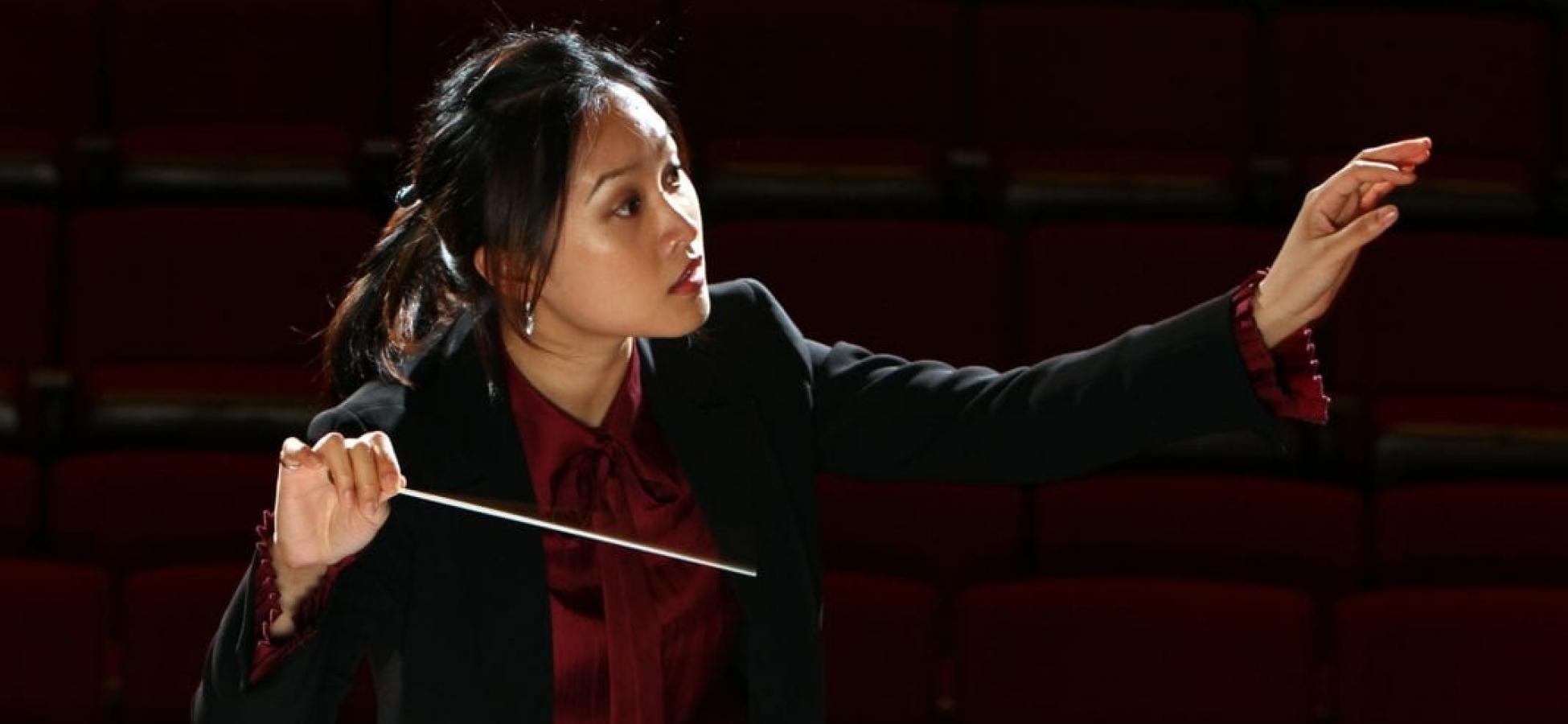 A person in formal attire focuses holds one hand out with a focused gaze in that direction, and the other hand holds a conductor's wand