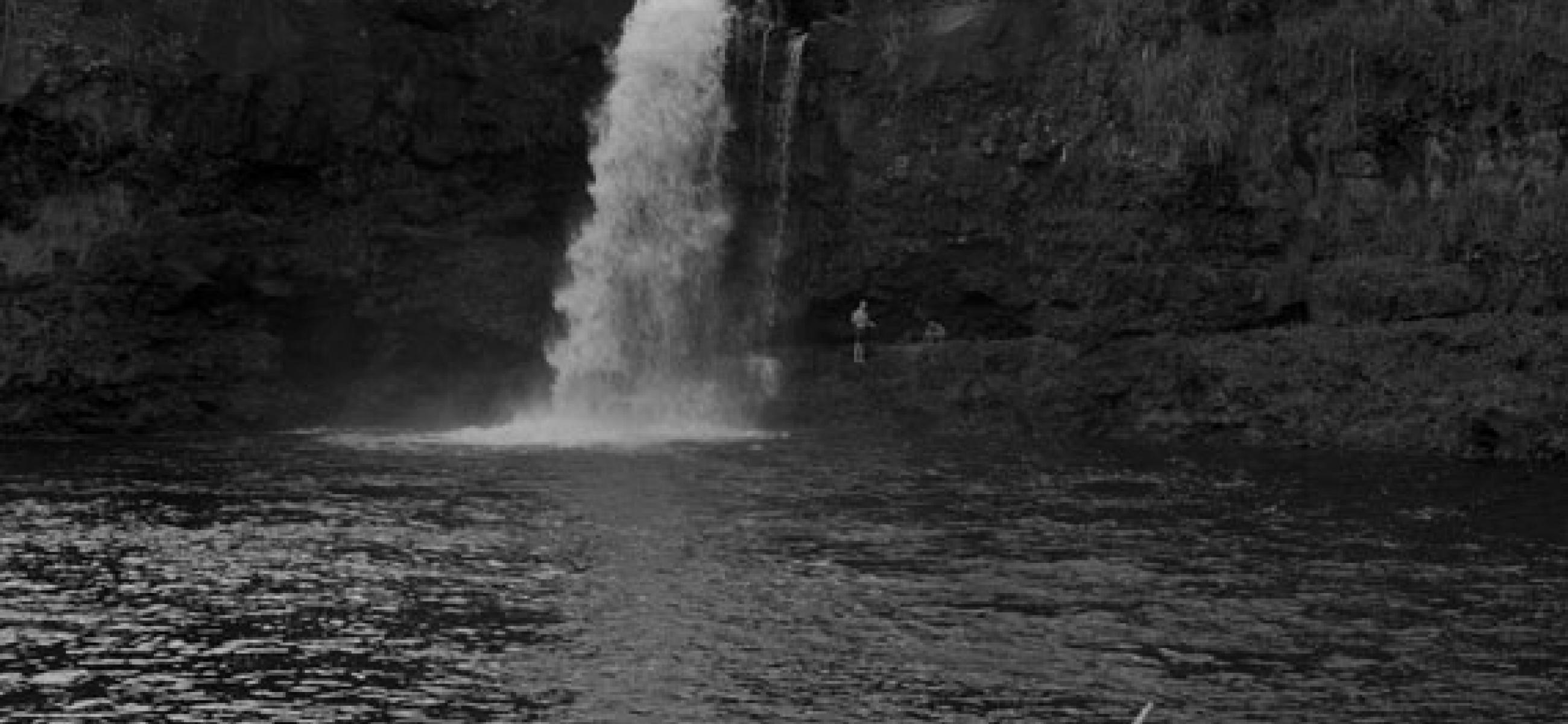 black and white photo of waterfall with pool and two people on shore in foreground