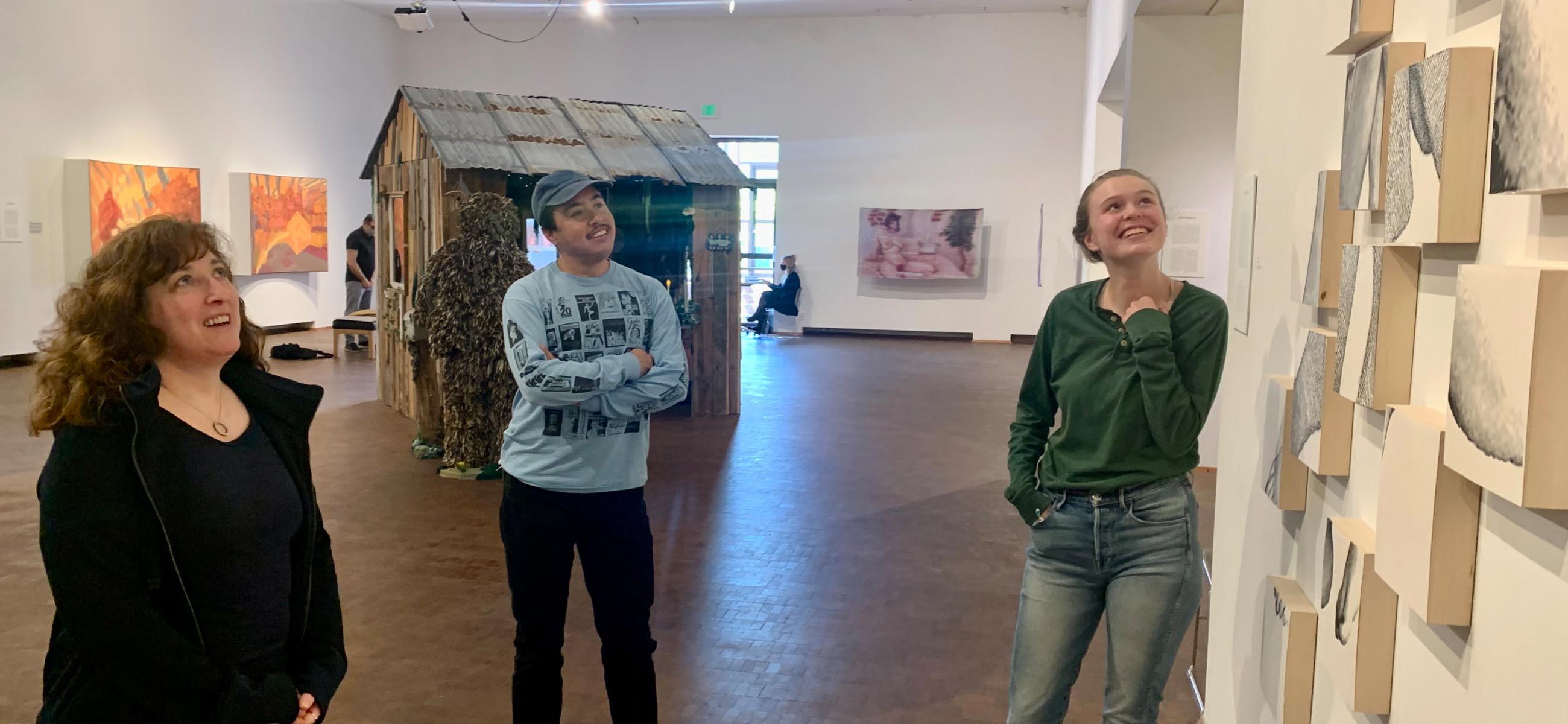 three smiling people look up at a group of artworks on the wall.