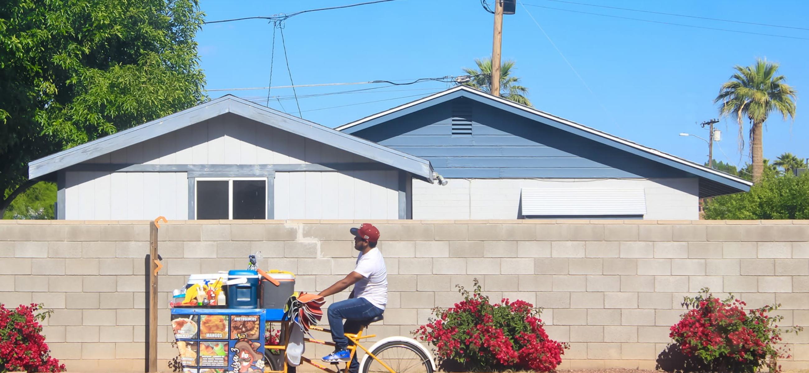 A person cycling past a house with a full cart on the front of the bike, almost as big as the bike