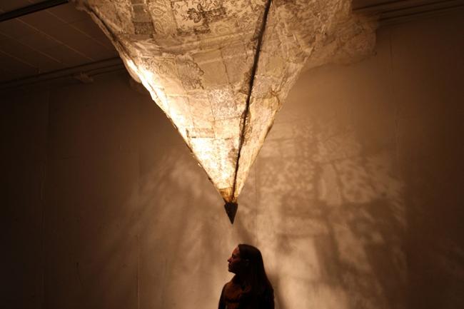 A silhouetted person stands looking up at the bottom point of an inner-lit, triangular hanging sculpture